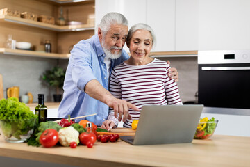 Happy Married Senior Couple Using Laptop While Cooking Healthy Food In Kitchen
