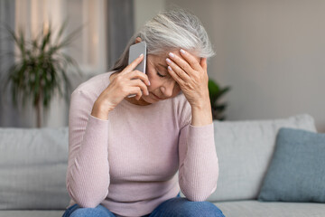 Depressed unhappy caucasian elderly gray-haired lady speaks by phone and holds her head with hand
