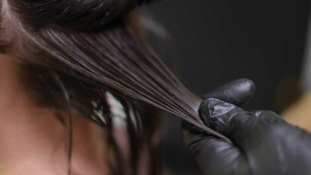 Close-up strand of brunette hair with hand of haidresser applying keratin straightening with infrared flat iron. Unrecognizable professional expert hairstylist treating damaged hair of client