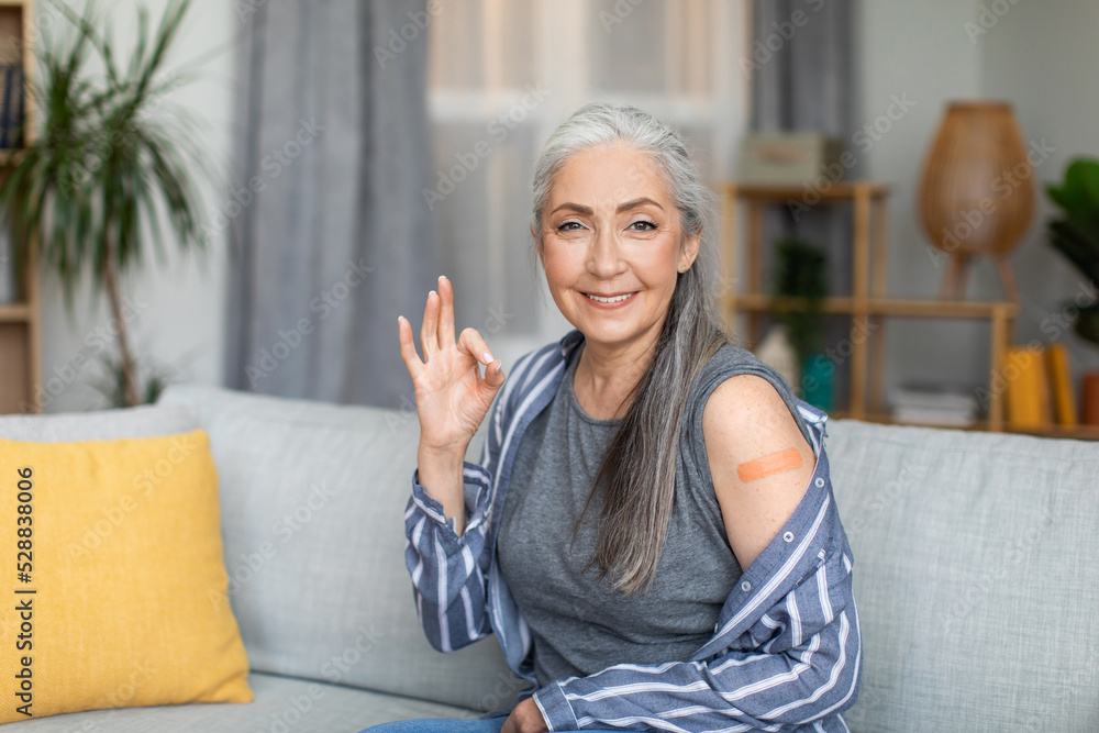 Wall mural smiling senior european gray-haired woman show ok sign with hand and shoulder with band aid after va