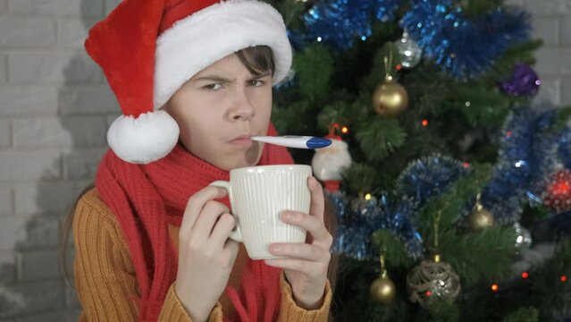 Ill girl at Christmas party. A sick teen girl in Santa hat stay with a mug of tea in the room.