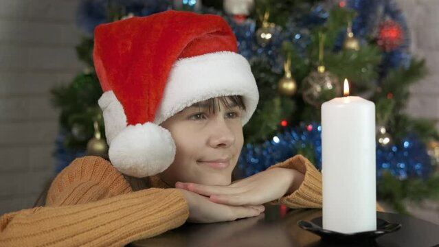Dreaming teen mood at Christmas eve. A view of dreaming teen in Santa red hat sits by the table with burning candle during magic New Year night.