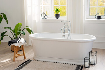 Fototapeta na wymiar Stylish modern bathroom interior. Horizontal view of an empty free-standing bathtub on a wooden floor in a bright room against the backdrop of a large window and houseplants. Soft selective focus.