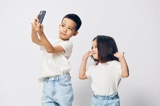 horizontal photo of happy, cute children, preschool age, brother and sister, in white shirts, taking a selfie on their smartphone, standing against a light background with empty space.
