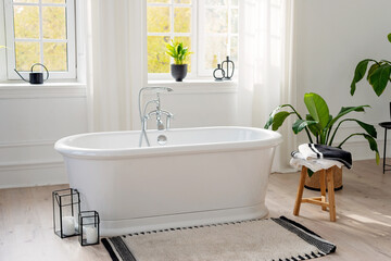 Fototapeta na wymiar Stylish modern bathroom interior. Horizontal view of an empty free-standing bathtub on a wooden floor in a bright room against the backdrop of a large window and houseplants. Soft selective focus.