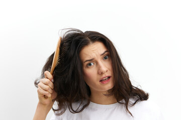 a sad, upset woman is trying to comb her long, dark, tangled hair with a wooden massage comb, standing in a white T-shirt on a white background and making a sad, funny face