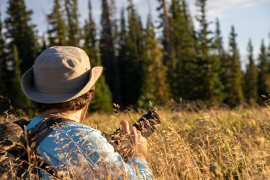 Photographer with DSLR wearing a outdoors adventure hat hiding in tall grass in Wyoming wilderness mountains looking for pictures to take