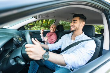 Cheerful millennial middle eastern guy driving car with lady in hijab, ride in car, enjoy music and dance