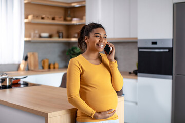 Happy pretty young pregnant african american woman touching belly and talking by phone