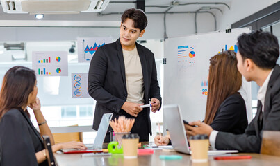 Asian handsome professional successful businessman presenter standing holding pen pointing presenting company graph chart paperwork document on glass board to male female colleagues in meeting room