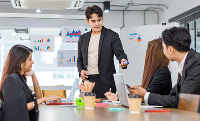 Asian handsome professional successful businessman presenter standing holding pen pointing presenting company graph chart paperwork document on glass board to male female colleagues in meeting room