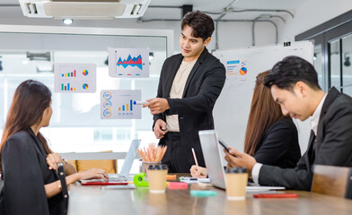 Asian handsome professional successful businessman presenter standing holding pen pointing presenting company graph chart paperwork document on glass board to male female colleagues in meeting room