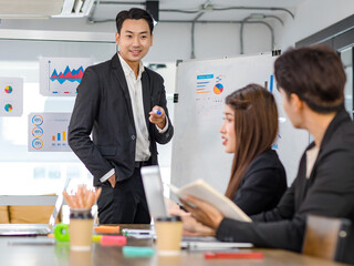 Asian handsome professional successful businessman presenter standing holding pen pointing presenting company graph chart paperwork document on glass board to male female colleagues in meeting room