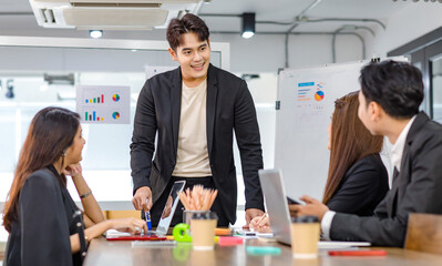 Asian handsome professional successful businessman presenter standing holding pen pointing presenting company graph chart paperwork document on glass board to male female colleagues in meeting room