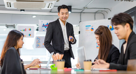 Asian handsome professional successful businessman presenter standing holding pen pointing presenting company graph chart paperwork document on glass board to male female colleagues in meeting room