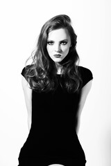 Fototapeta premium Beautiful woman with long hair studio portrait in white background. Model sitting on chair with hands placed behind her back and wearing black sexy dress. Smokey make-up. Black and white image