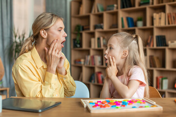 Professional woman speech therapist studying together with pretty little girl, learning practice...