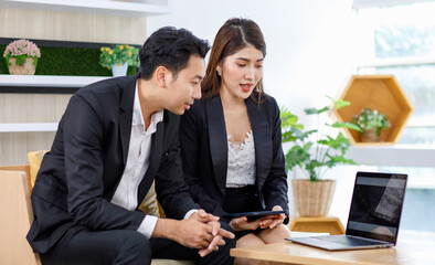 Asian young beautiful professional successful businesswoman in formal suit sitting smiling holding tablet pointing showing information data from laptop notebook computer to male businessman colleague