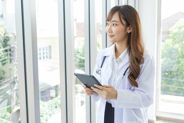 Beautiful and cheerful good mood young lady asian doctor dressed uniform smiling arm stethoscope while using tablet on lobby hospital background..