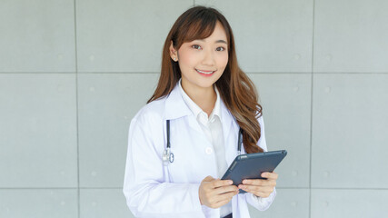 Beautiful and cheerful good mood young lady asian doctor dressed uniform smiling arm stethoscope while using tablet on lobby hospital background..