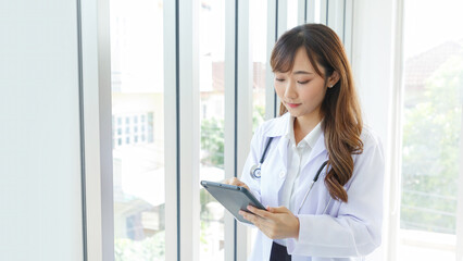 Beautiful and cheerful good mood young lady asian doctor dressed uniform smiling arm stethoscope while using tablet on lobby hospital background...