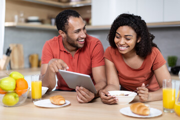Laughing young african american couple have breakfast, enjoy chatting, watching video in tablet