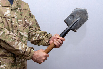 A soldier in an American military uniform holds a sapper shovel in his hands on a light background.