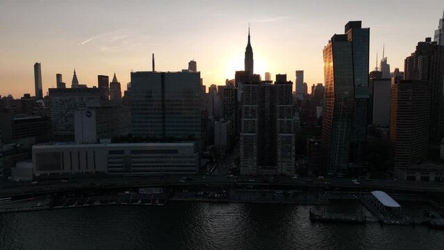 An aerial view of Manhattan over the East River in NY at the Manhattan Henge sunset of 2022. The camera truck left viewing the NYC skyline in silhouette, with sunlight shining between the buildings.