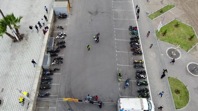 Drone video of a motorcycle competition. Many motorcycles parked as two people practice their wheelies and turns. Drone hovers and slowly tilts camera up. Located in Chorrillos, Lima, Peru.