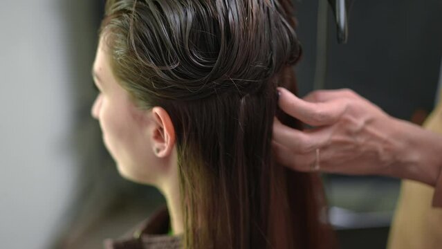 Hairdresser with hair dryer drying long brunette laminated hair of client in slow motion. Close-up Caucasian lady sitting in hair salon with unrecognizable hairstylist doing keratin aftercare
