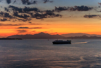 Cargo ship sails through calm seas off coast with distant sunset