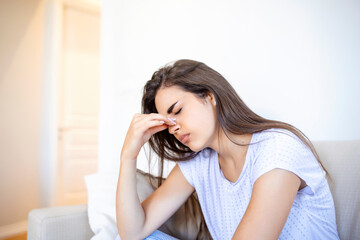 Portrait of an attractive woman sitting on a sofa at home with a headache, feeling pain and with an expression of being unwell. Upset woman sitting on couch feeling strong headache migraine.