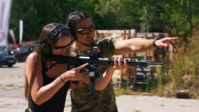 Man and woman wearing safety headphones and goggles practicing using submachine gun under supervision of instructor. Firearms training at outdoor shooting range, Horizontal shot. High quality 4k