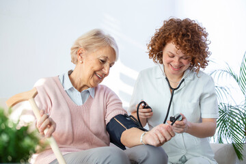 Happy senior woman having her blood pressure measured in a nursing home by her caregiver. Happy nurse measuring blood pressure of a senior woman in living room