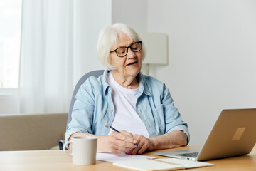 Fototapeta premium a happy elderly woman is sitting at her desk at home and holding an online conference on a laptop, listening attentively to the information
