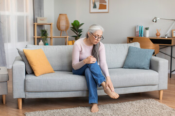 Sad caucasian elderly gray-haired woman in glasses sit on sofa touches leg, suffering from paine