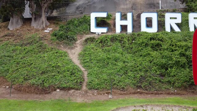 Drone video of a red sign reading "Peru" and behind it on a green hill another sign reading "Chorrillos" the name of the district in which this was recorded. Drone slowly flies left to right.