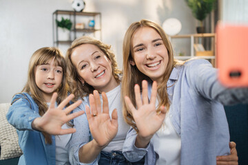 Attractive beautiful two people two young lady girls sisters and their mother using mobile phone taking photo, picture or having video call while sitting at home on sofa, waving hands.