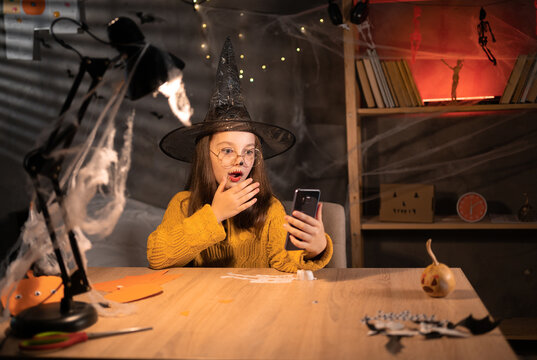 Portrait Of Scared Girl In Witch Hat Looking At Mobile Phone Seeing Bad News, Photos Or Message. Worried Teen Grabbing Head At Darkness Room On Halloween. Human Reaction