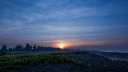 Obraz premium The golden orange sun is rising through the trees behind the sand under the bush at Suruchi beach in Vasai, breaking through the cloud lines of the blue sky.