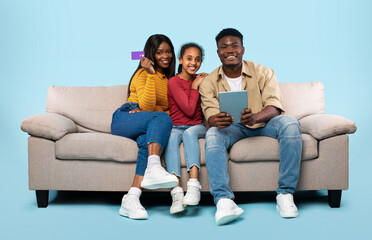 Fototapeta na wymiar Happy black family of three using digital tablet and credit card for online shopping, sitting on couch, blue background