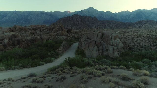 Drone pushes over a massive boulder toward Mt Whitney and the Sierra Nevada mountain range in Alabama Hills California at dusk, as an SUV drives toward camera