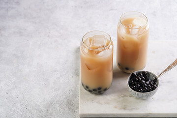 Two glasses with black tea, milk, ice cubes and cooked tapioca pearls for trendy bubble boba ice tea, two small grey ceramic bowls on marble board on pastel grey background