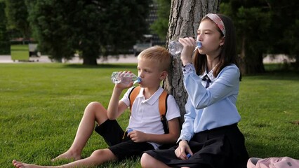 Schoolchildren with backpacks are sitting on the grass and drinking water.