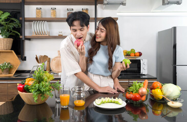 Asian couple spend time together in the kitchen. Young man in casual cloth bites the delicious red apple in his girlfriend hand. The counter full of various kinds of vegetables and fruit juice.