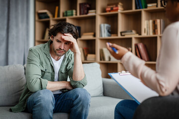 Unhappy depressed young european male patient listens to black woman psychologist in clinic interior