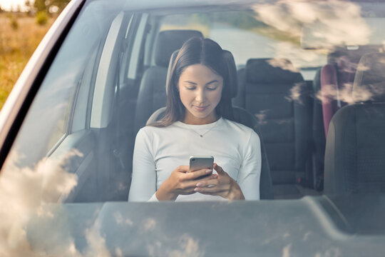 Portrait of beautiful dark haired woman wearing white shirt sitting in car and texting a message, holding mobile phone and checking social networks while waiting a driver.