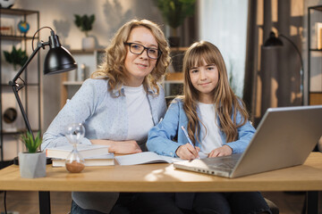 Portrait of joyful small daughter and her middle age mother doing homework and looking at camera. Young mom and her cute girl happy to make right tasks, sitting at home.