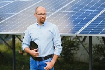 Portrait of male engineer with tablet in his hands near the solar panels station, wearing helmet at sunny day. Green ecological power energy generation.