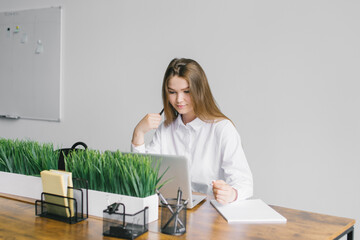 A cute girl is sitting at a wooden table with greenery in the office working with a laptop